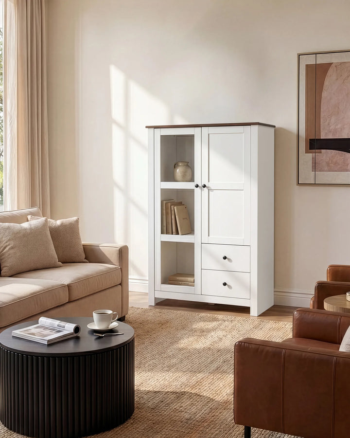 A tall white cabinet with two glass-front upper doors and two lower drawers, styled in a bright living room next to a beige sofa and round black coffee table.