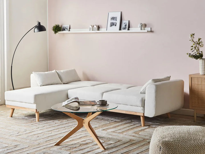 A modern light beige sectional sofa with wooden legs in a minimalist living room setting, styled with a glass-top coffee table, a black floor lamp, and a pink wall.