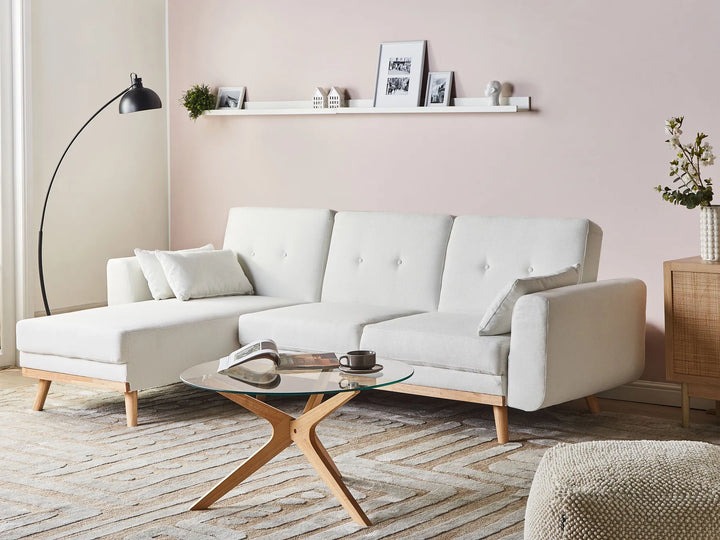 A modern white sectional sofa with light wood legs in a styled living room setting, featuring a glass-top coffee table, a black floor lamp, and a pink wall background.