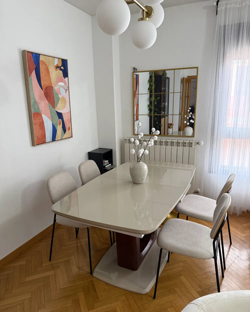 A modern dining room setup featuring a glossy white rectangular dining table with a dark brown pedestal base, surrounded by six light beige upholstered dining chairs with black metal legs. The room has white walls, a colorful abstract art print, a gold-framed mirror, and a white globe chandelier.