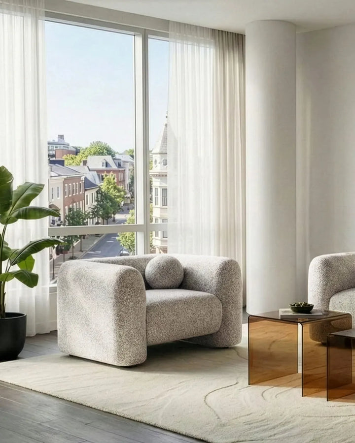 A modern light beige bouclé armchair with a round matching pillow, styled in a bright, contemporary living room with a large window, sheer curtains, and a glass side table.