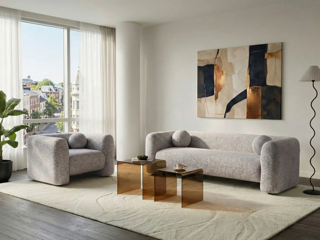 A modern living room featuring a light gray textured sofa and matching armchair on a large cream rug, with a gold mirrored coffee table in front. A large abstract painting hangs on the wall above the sofa, and a window with sheer curtains is visible on the left.