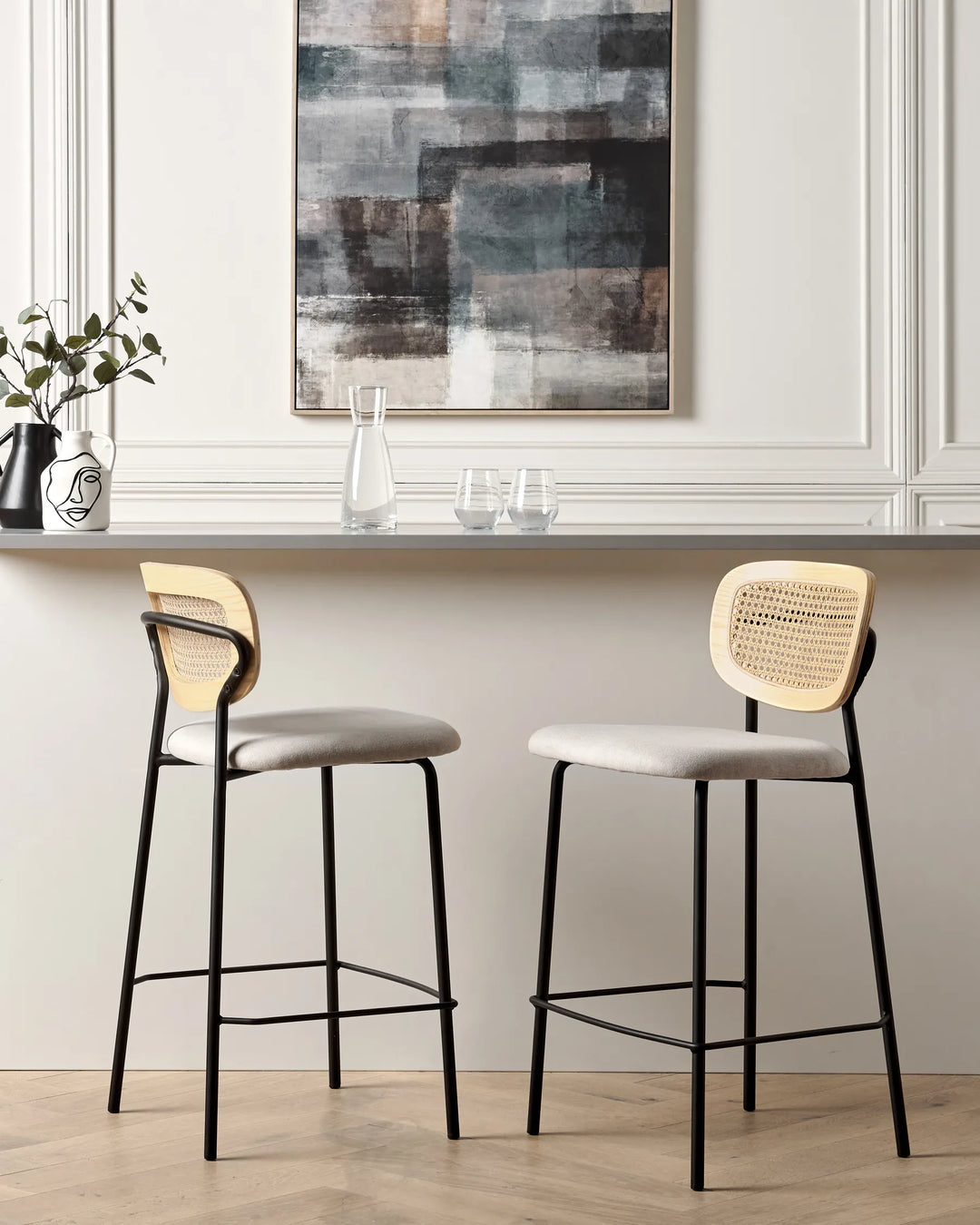 Two modern bar stools with light beige woven cane backs and light gray upholstered seats, set in a bright interior with a white paneled wall, a gray counter, and a framed abstract painting.