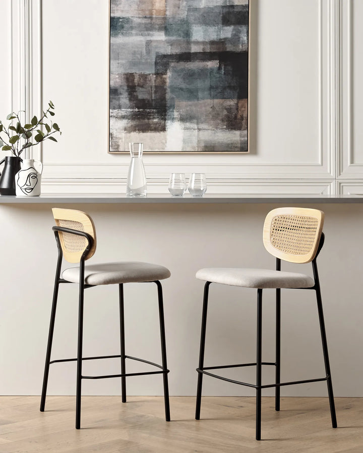 Two modern bar stools with light beige woven cane backs and light gray upholstered seats, set in a bright interior with a white paneled wall, a gray counter, and a framed abstract painting.
