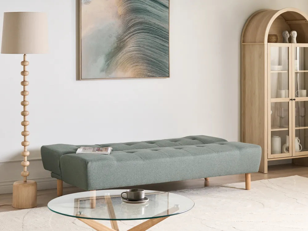 A tufted daybed in a muted sage green fabric with light wood legs, styled in a bright, modern living room next to a wooden floor lamp and a glass coffee table.