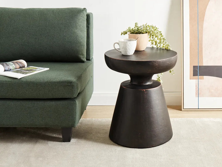 A dark brown wooden accent side table with a tiered pedestal base, styled in a living room next to a green armchair with a coffee mug and potted plant on top.