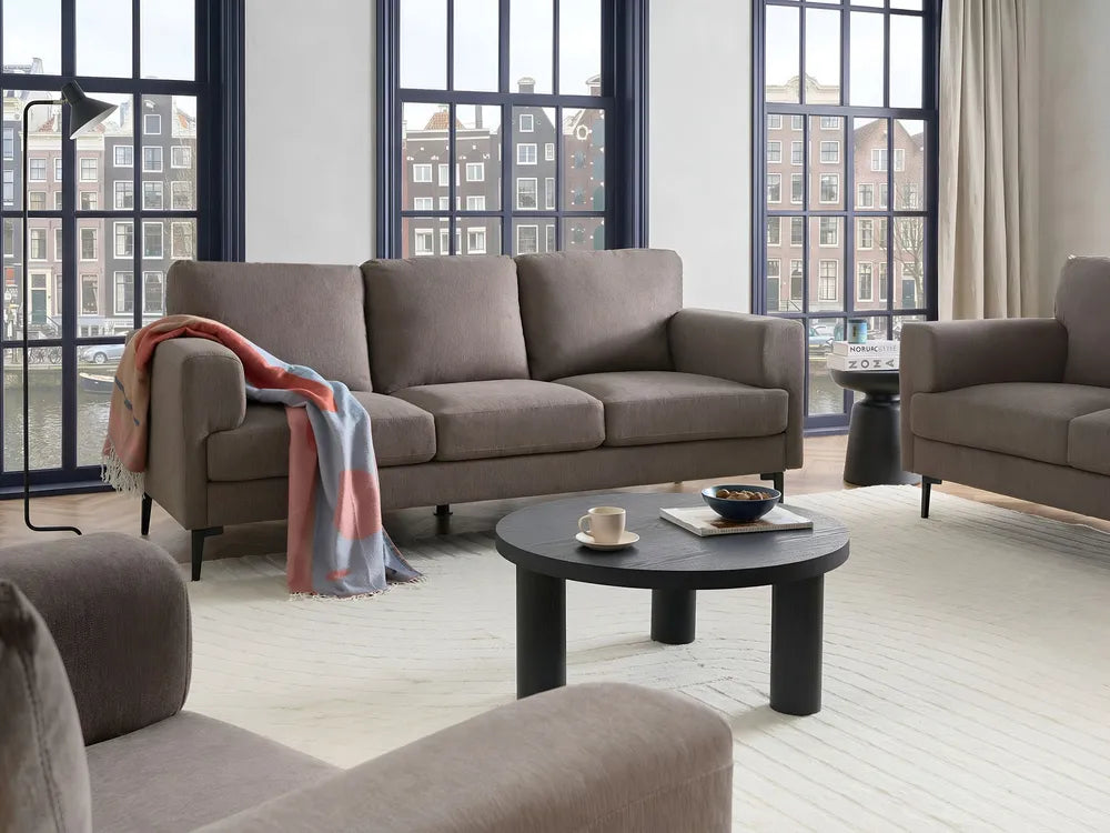 A modern living room scene featuring a taupe three-seater sofa, a matching armchair, a round black coffee table, and a draped throw blanket, set against large multi-pane windows with a city view.