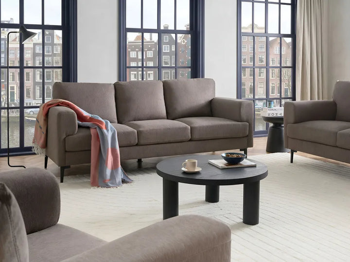 A modern living room scene featuring a taupe three-seater sofa, a matching armchair, a round black coffee table, and a draped throw blanket, set against large multi-pane windows with a city view.