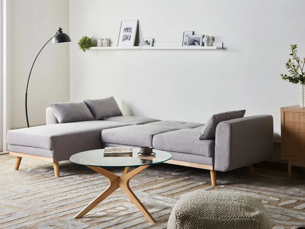 A modern gray fabric sectional sofa with light wood legs, styled in a bright minimalist living room with a glass-top coffee table, floor lamp, and wall shelf.