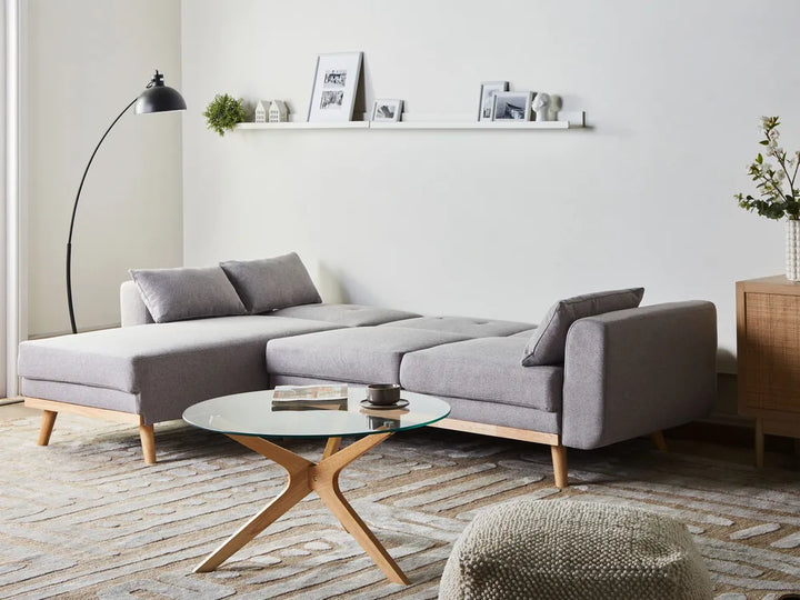 A modern gray fabric sectional sofa with light wood legs, styled in a bright minimalist living room with a glass-top coffee table, floor lamp, and wall shelf.