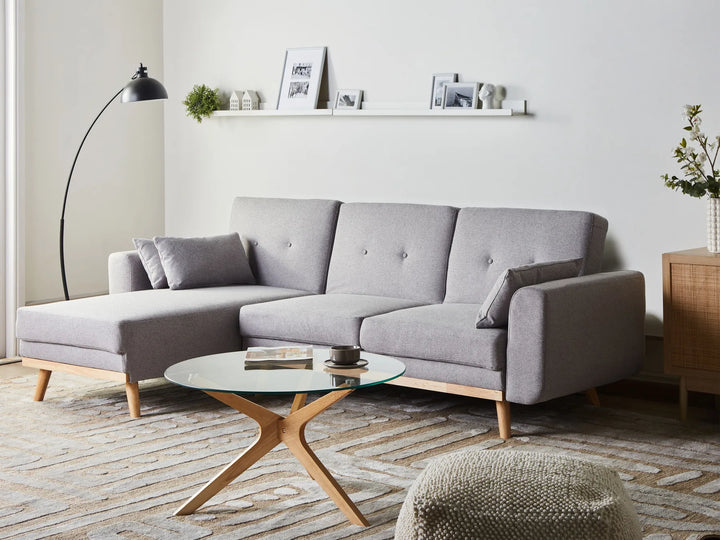 A light gray fabric sectional sofa with a chaise lounge and light wood legs, styled in a modern living room setting with a glass-top coffee table, a black floor lamp, and a neutral rug.