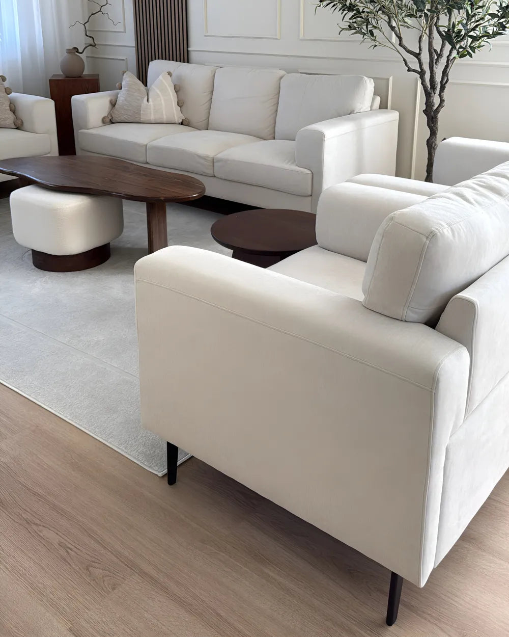 A modern living room setup featuring a cream-colored upholstered armchair in the foreground, a matching cream sofa in the background, a dark wood coffee table, and a light gray rug on a wood-look floor.