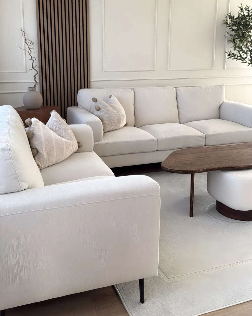 A modern living room setup featuring a light beige upholstered sectional sofa and matching armchair with decorative pillows, a dark wood oval coffee table, a white cylindrical side table, and a light rug on a wood floor.