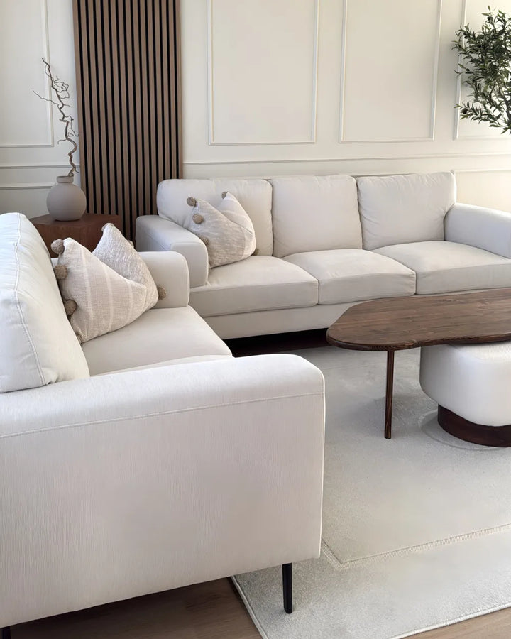 A modern living room setup featuring a light beige upholstered sectional sofa and matching armchair with decorative pillows, a dark wood oval coffee table, a white cylindrical side table, and a light rug on a wood floor.