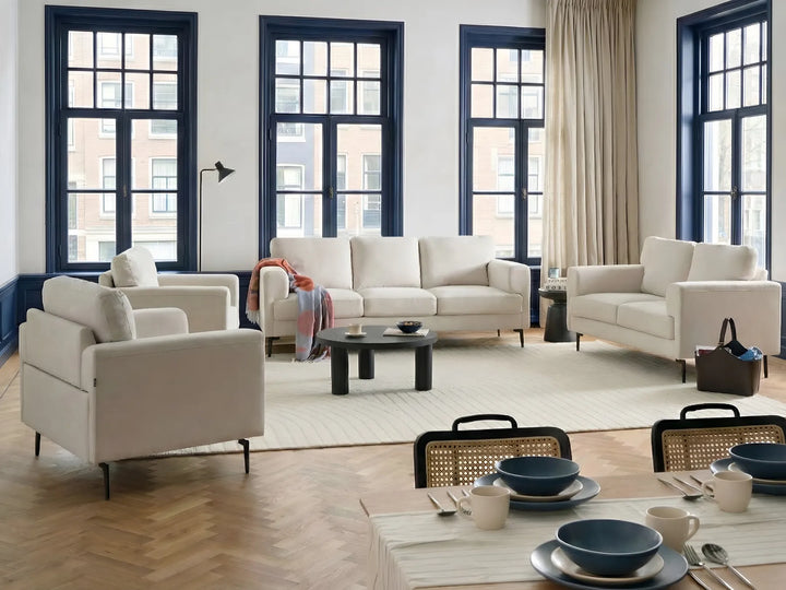 A styled living room featuring a cream-colored sofa, matching armchair, and loveseat arranged around a round black coffee table on a large off-white rug. The room has herringbone wood floors, tall windows with dark blue frames, and beige curtains.