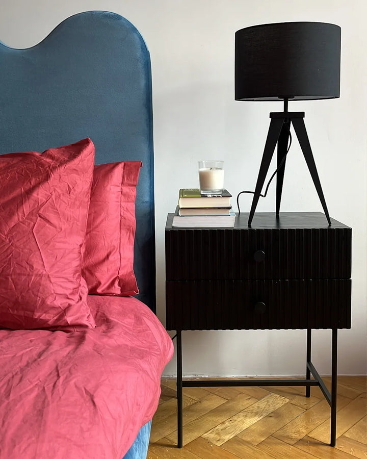 A black two-drawer nightstand with a ribbed front and slim metal legs, styled in a bedroom next to a bed with a blue headboard and pink bedding. A black tripod table lamp sits on top of the nightstand next to a stack of books.