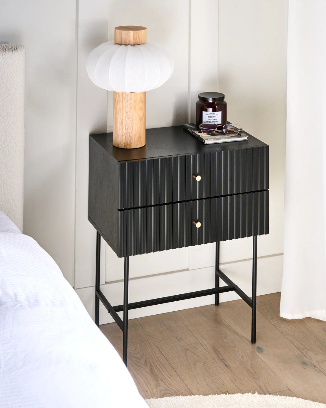 Black nightstand with two drawers, a lamp, and a jar on a wooden floor.