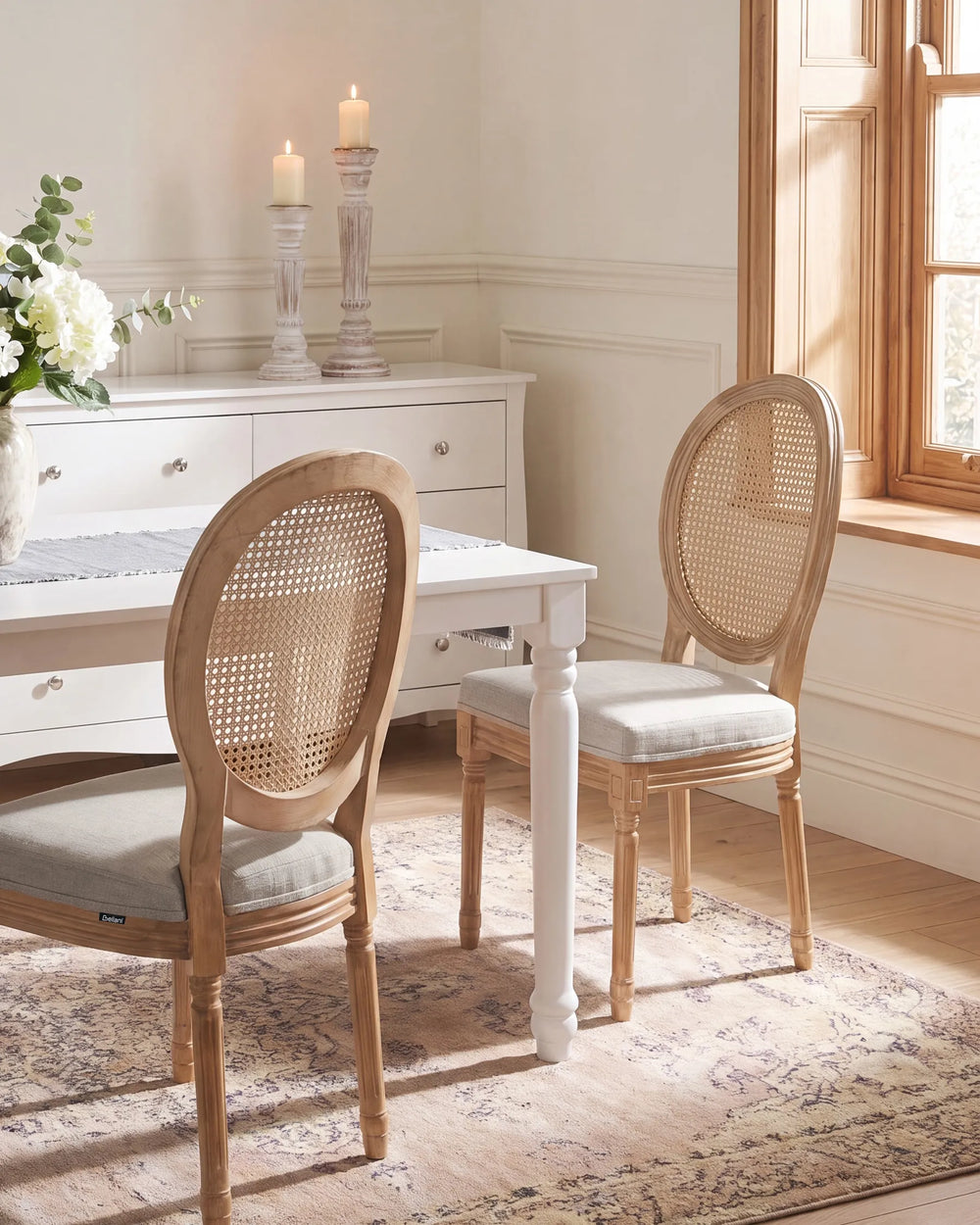 A dining room scene featuring two light wood dining chairs with woven cane backs and light gray upholstered seats, set around a white table on a patterned rug.