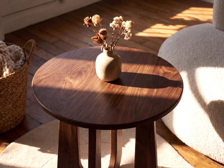 A round dark wood side table with a small beige ceramic vase holding dried flowers, placed in a sunlit room with a woven basket, wood floor, and cream upholstered chair visible.
