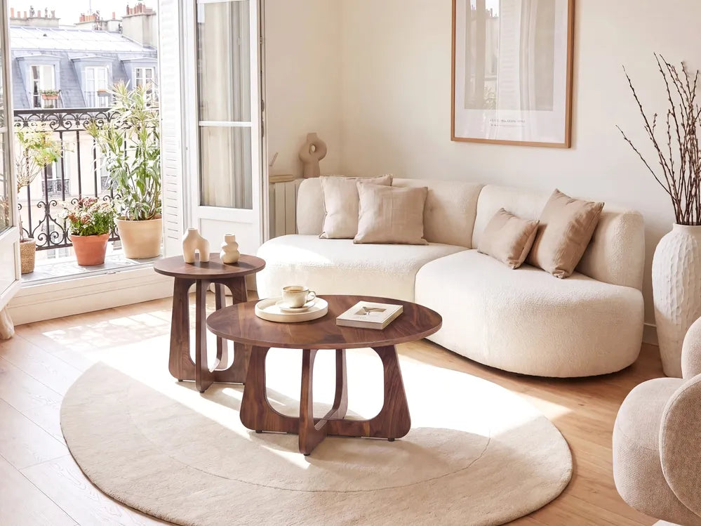 A modern living room featuring a cream-colored curved sectional sofa, two dark brown wooden nesting coffee tables, a round beige rug, and a large window overlooking a balcony with potted plants.