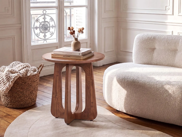 A round wooden side table with a sculptural curved legs design, styled in a bright, modern living room next to a cream bouclé sofa and a woven basket.