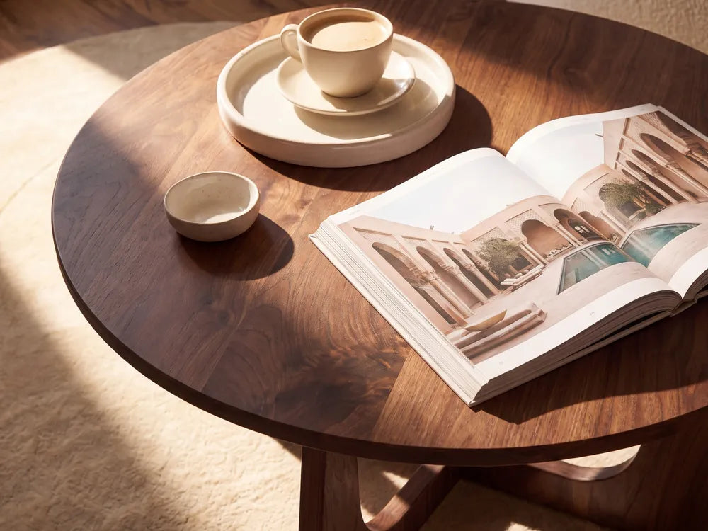 A round dark wood table styled with a ceramic cup and saucer on a tray, a small bowl, and an open architectural magazine. The scene is lit by warm, directional sunlight casting soft shadows on a light beige rug.