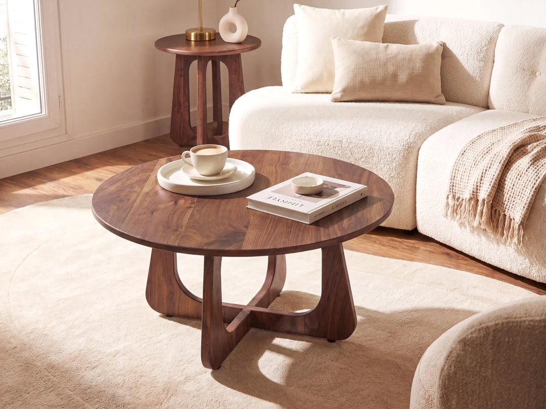 A round wooden coffee table with a dark walnut finish and sculptural curved legs, styled in a bright, modern living room next to a cream bouclé sofa.