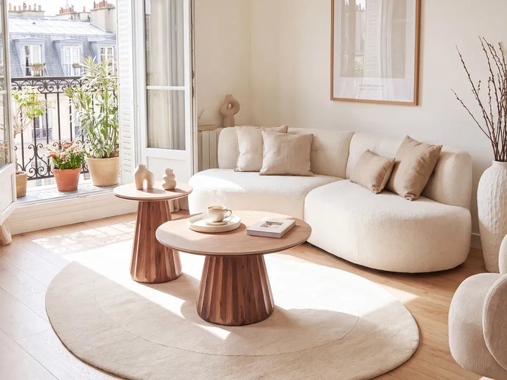 A modern living room featuring a curved cream bouclé sectional sofa, two round wooden coffee tables, a large cream circular rug, and a bright window with a balcony view.