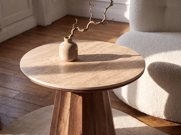 A round wooden side table with a light natural stone top and a dark wood pedestal base, styled with a small ceramic vase holding a dried branch in a sunlit room next to a cream boucle chair.
