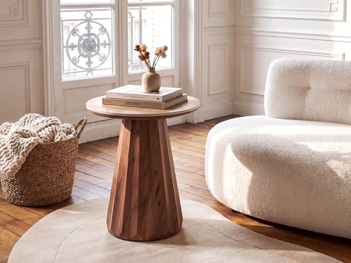 A round wooden side table with a tapered, faceted pedestal base, styled in a bright, modern living room next to a cream bouclé sofa and a woven basket.