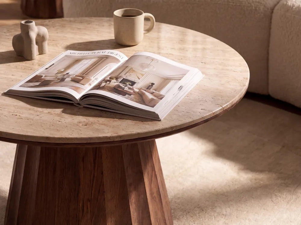 An open magazine lies on a round light stone-top coffee table in a warm, sunlit living room. A beige mug sits behind it, with a cream sofa visible in the background.