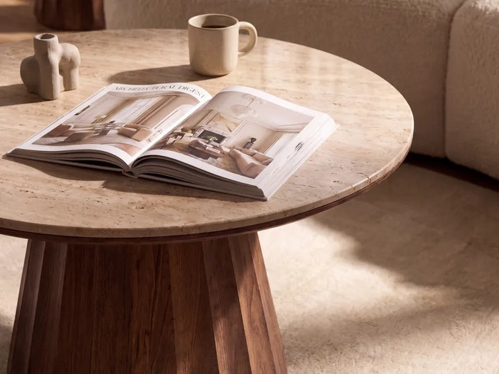 An open magazine lies on a round light stone-top coffee table in a warm, sunlit living room. A beige mug sits behind it, with a cream sofa visible in the background.