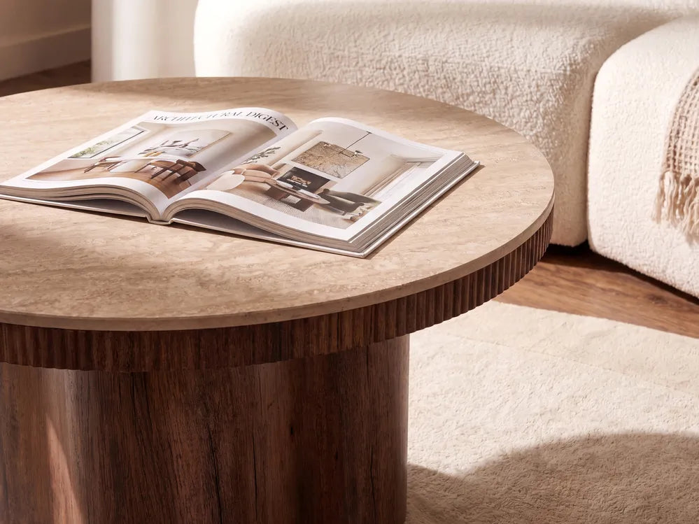 A round wooden coffee table with a textured light beige top and a darker brown fluted pedestal base, styled in a living room with an open magazine on top and a cream-colored sofa in the background.