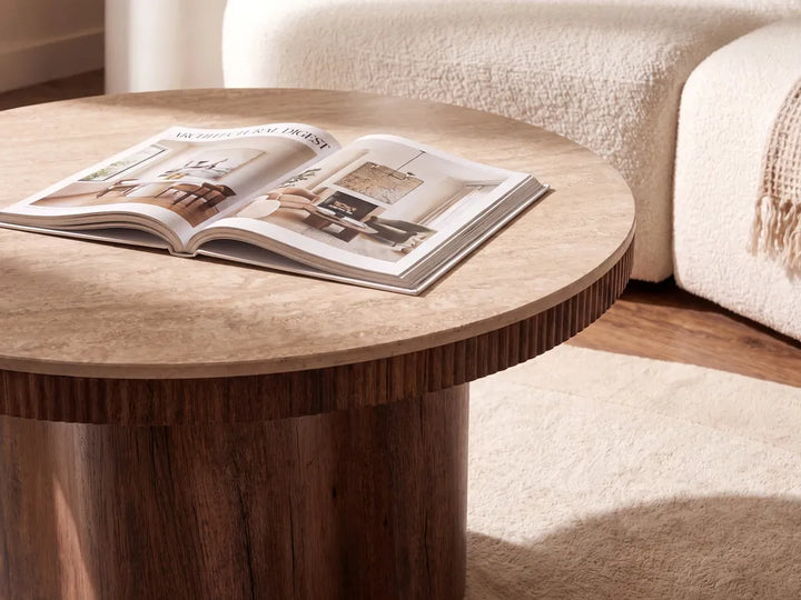 A round wooden coffee table with a textured light beige top and a darker brown fluted pedestal base, styled in a living room with an open magazine on top and a cream-colored sofa in the background.