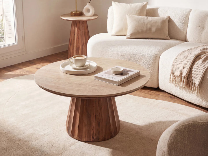 A round coffee table with a light wood top and a sculptural fluted pedestal base, styled in a bright, modern living room next to a cream bouclé sofa.