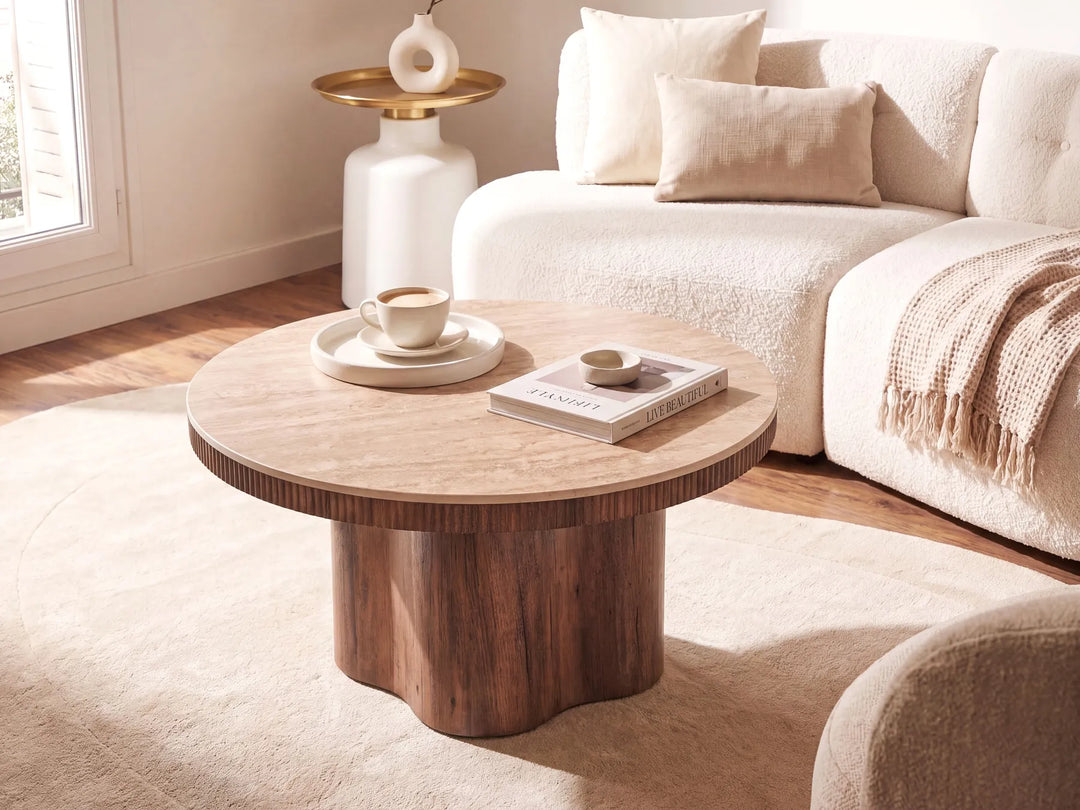 A round wooden coffee table with a chunky pedestal base and a flared top edge, styled in a bright, modern living room next to a cream bouclé sofa.