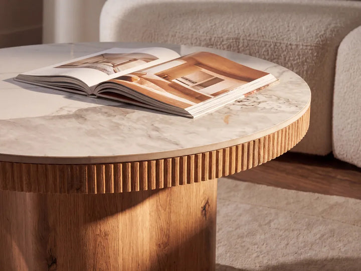 A round coffee table with a white marble top and a fluted light wood pedestal base, shown in a warm interior setting with an open magazine on the surface.
