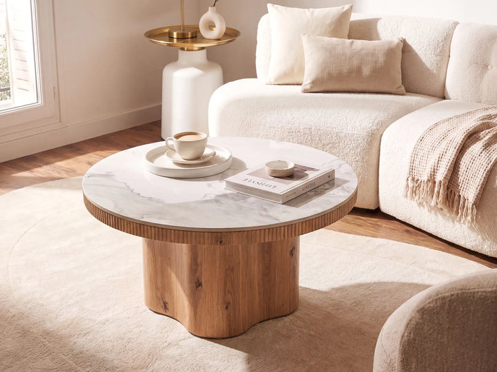A round coffee table with a white marble top and a sculptural wavy wooden base, styled in a bright, modern living room next to a cream bouclé sofa.