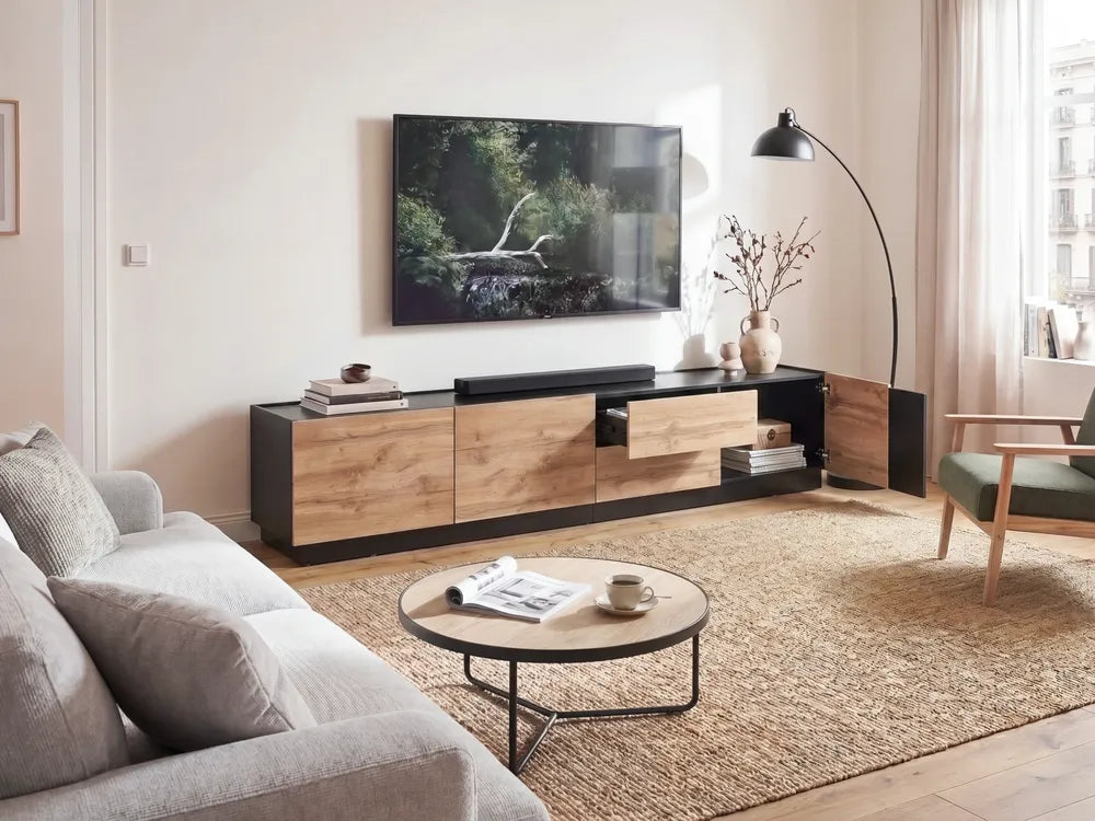 A modern living room setup featuring a low wooden TV console with black accents, a wall-mounted flat-screen TV, a round coffee table, a textured rug, and a floor lamp.