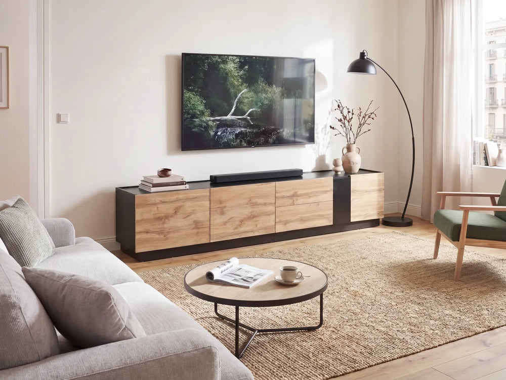 A modern living room setup featuring a low wooden TV console with black side panels, a wall-mounted flat-screen TV, a round coffee table, a beige rug, and a black floor lamp.