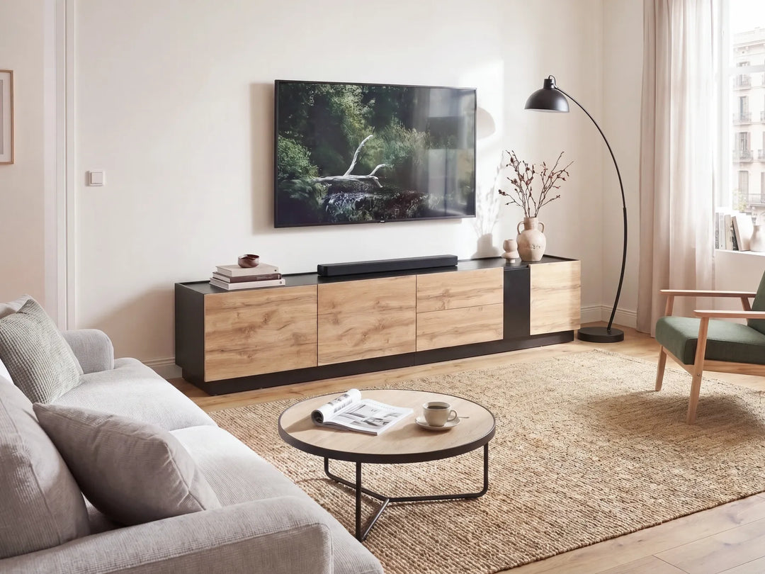 A modern living room setup featuring a low wooden TV console with black side panels, a wall-mounted flat-screen TV, a round coffee table, a beige rug, and a black floor lamp.