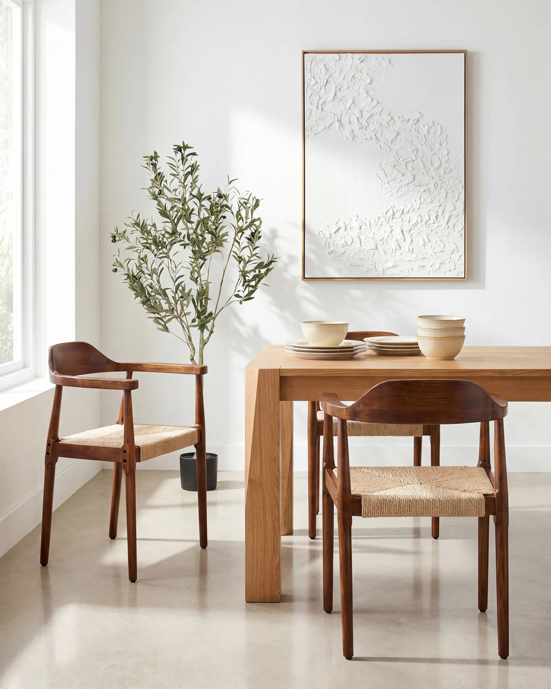 A mid-century modern dining room scene featuring a light oak wood table and two matching wooden chairs with woven beige seat cushions, styled with ceramic bowls and a potted olive tree against a white wall with a framed abstract print.