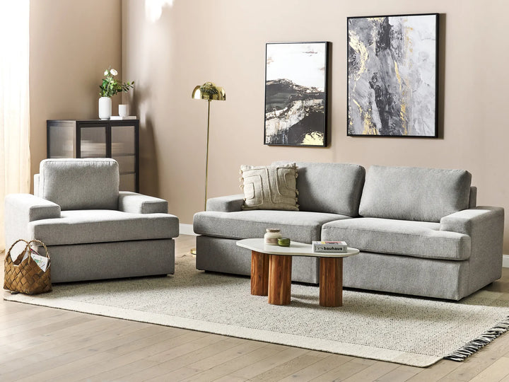 A modern living room set featuring a light gray upholstered three-seater sofa and matching armchair, styled with a low wooden-topped coffee table, textured rug, and framed wall art.