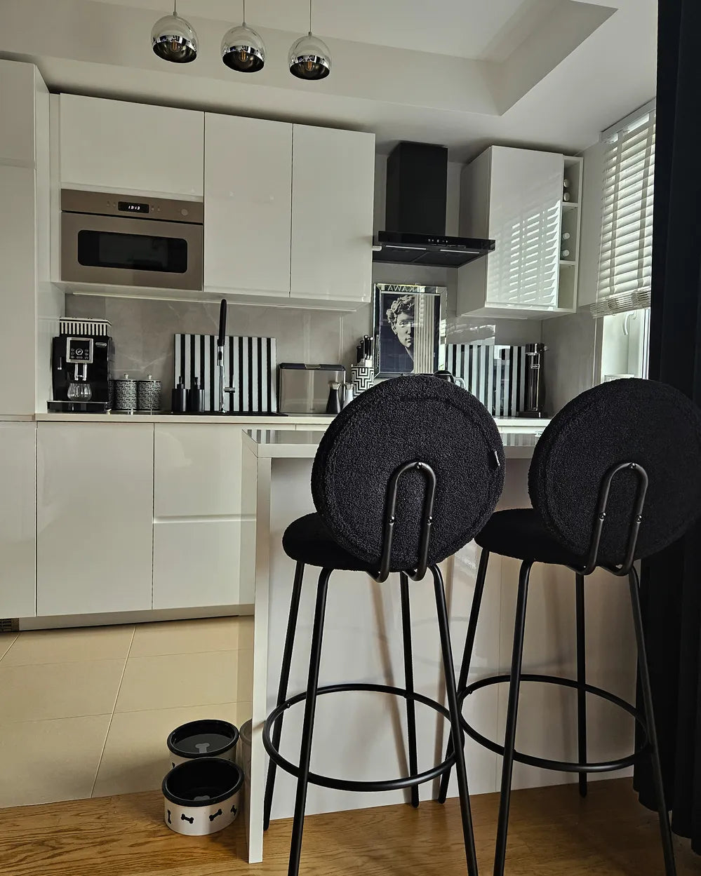 A modern kitchen interior with white cabinets, a stainless steel microwave, black-and-white striped accents, and two black bar stools in the foreground.
