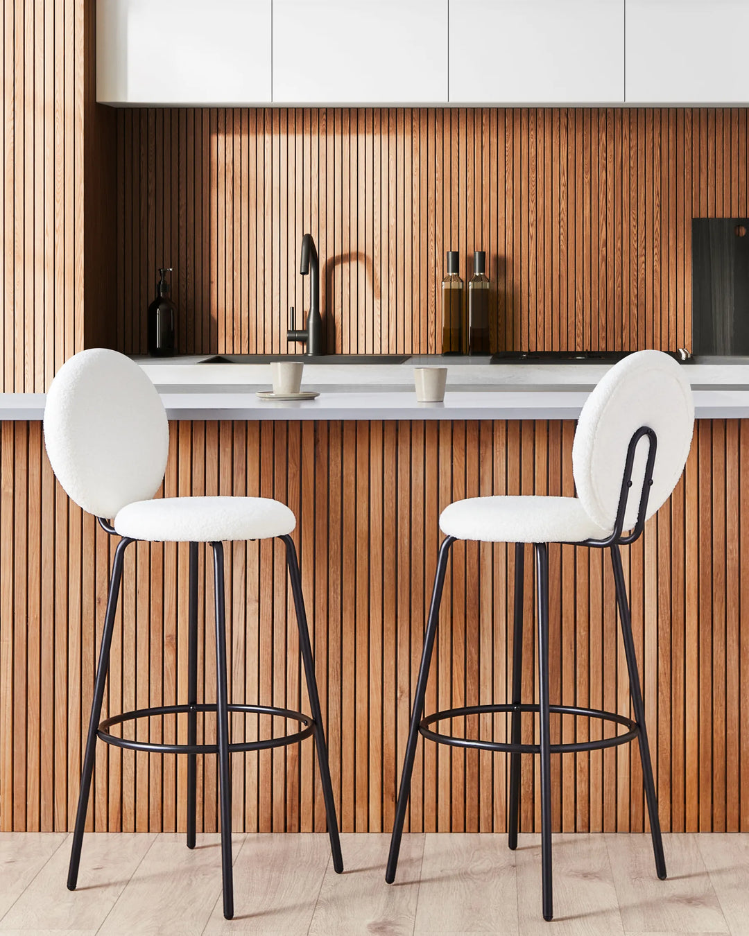 Two modern bar stools with white upholstered seats and backs and black metal frames, set in front of a wood slat kitchen backsplash.