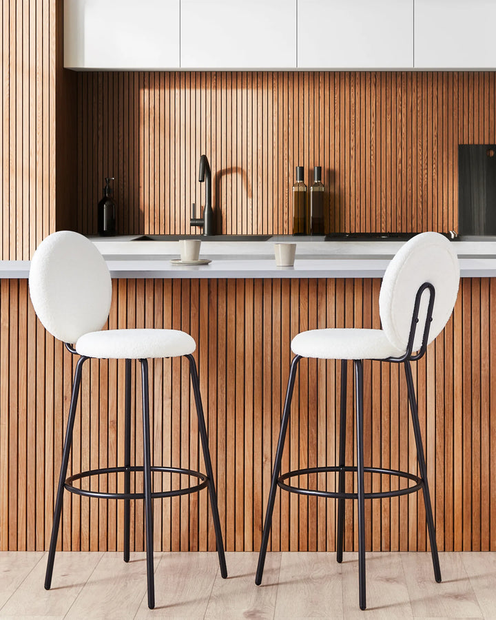 Two modern bar stools with white upholstered seats and backs and black metal frames, set in front of a wood slat kitchen backsplash.