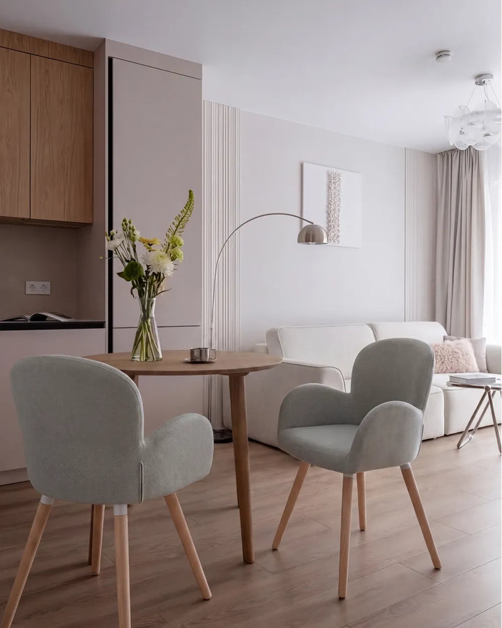 A modern dining area in a bright, minimalist apartment featuring a light wood dining table and two light gray upholstered chairs with wooden legs, set against a neutral living room background with a white sofa and beige curtains.