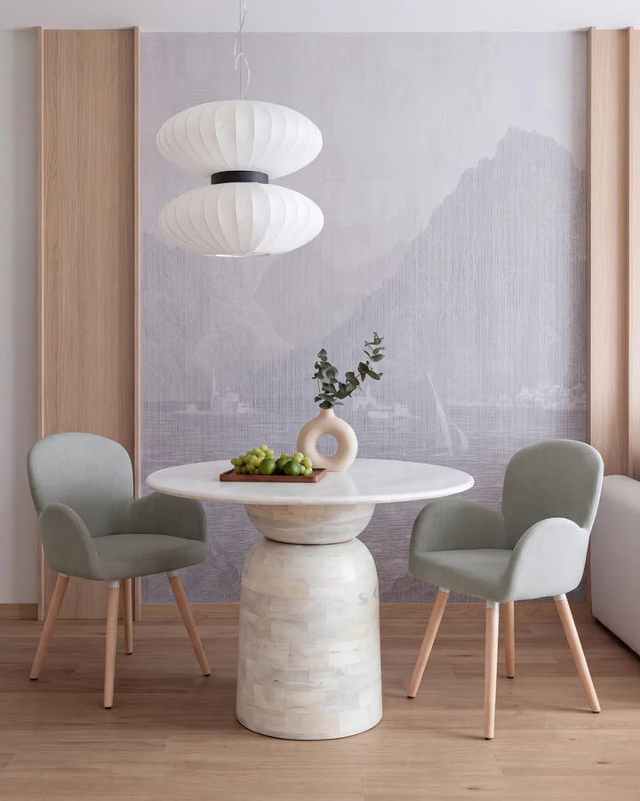 A modern dining room setup featuring a round white marble-top pedestal table, two light sage green upholstered chairs with light wood legs, a double-tier white paper lantern pendant light, and a gray mountain mural on the back wall.