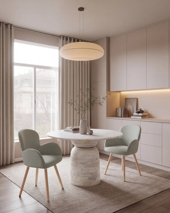 A modern dining room scene featuring a round white stone pedestal table, a matching round pendant light, two pale sage green upholstered chairs with light wood legs, and a neutral beige rug in a bright, contemporary interior.