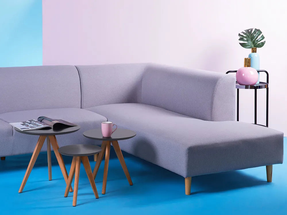 A modern light purple sectional sofa with a chaise lounge, styled in a minimalist living room setting with three small wooden-topped side tables, a pink mug, and a black side table holding a plant against a pastel pink and blue background.